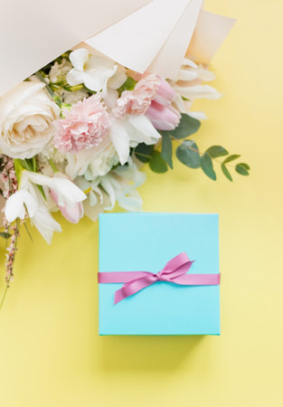 Beautiful bouquet with roses, carnations, iris and eucalyptus and blue gift box with pink ribbon on a yellow background, copy space. Mothers day, 8 march, holiday gift conceptの写真素材