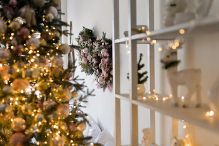 Pink and golden Christmas decorations on a Christmas tree with lights, wreath on the white wall and white wooden shelving with lights and decorの写真素材