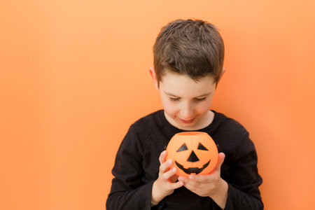 Caucasian boy in black clothes holding Halloween pumpkin Jack on an orange background, copy space. Halloween mockup with caucasian boyの写真素材