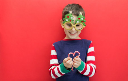 Caucasian boy in funny glasses holding tasty candy canes on a red background, copy spaceの写真素材