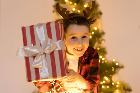 Boy in reindeer horns holding gift box near Christmas treeの写真素材