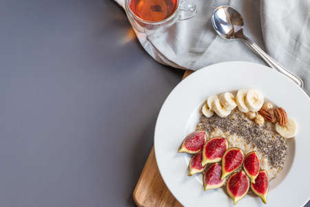 a bowl with breakfast oatmeal porridge with superfood chia seeds, fresh figs and banana on a wooden board on grey background , served with black tea, , shot from aboveの写真素材