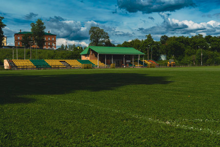 A soccer field with green grass, empty stands, and a green-roofed shelter under a cloudy sky.の写真素材