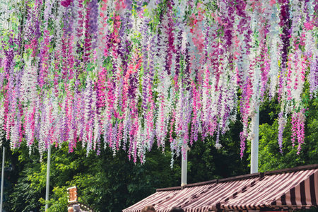 Colorful artificial flowers hanging from a canopy at an outdoor festival, with green trees in the background.の写真素材