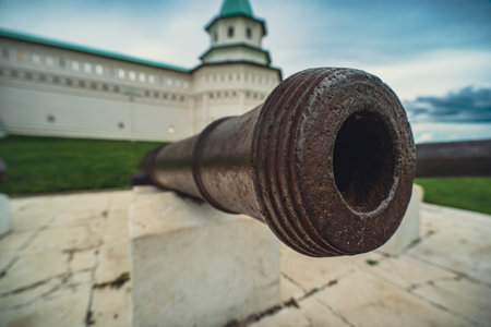old, rusty cannon aimed directly at the camera, with a historical building in the background. Appearance of the cannon, set against a cloudy sky, historical significance and the passage of time.の写真素材
