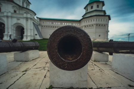 old, rusty cannon aimed directly at the camera, with a historical building in the background. Appearance of the cannon, set against a cloudy sky, historical significance and the passage of time.の写真素材