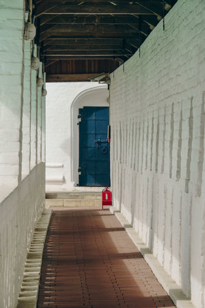 A narrow historic corridor with white walls, arched doorways, and a wooden ceiling. The hallway leads to a large, black metal door, creating a sense of depth and antiquity.の写真素材