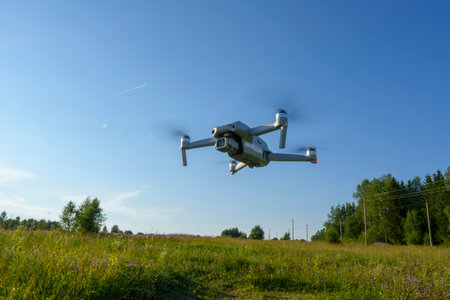 A quadcopter drone flying low over a grassy field on a clear, sunny day. The sky is blue and the landscape is lush with greenery, illustrating modern technology in a natural setting.の写真素材