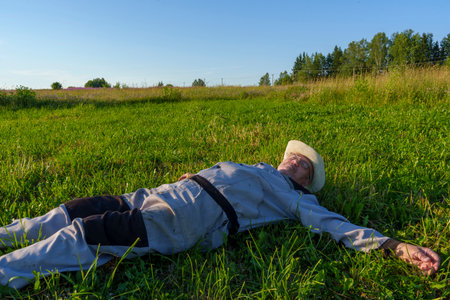An elderly man wearing a hat and overalls lies peacefully on his back in a grassy field under a clear blue sky. The scene exudes tranquility and relaxation in a rural setting.の写真素材