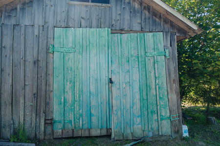 An old wooden barn with weathered green doors, situated in a rural area and surrounded by trees on a...