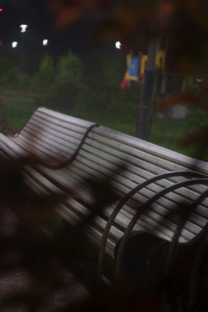 A close-up view of an empty park bench at night, with a blurred background of lights and foliage, creating a tranquil and solitary atmosphere.の写真素材