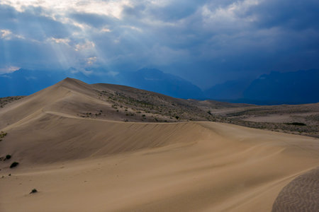 Expansive desert dunes stretch under a dramatic sky with sunbeams breaking through the clouds, creating a serene and awe-inspiring landscape.の写真素材