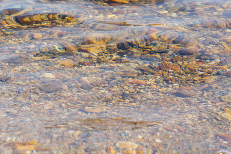 A close-up view of clear water gently ripping over colorful pebbles at the bottom of a lake,...