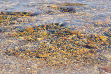 A close-up view of clear water gently ripping over colorful pebbles at the bottom of a lake, highlighting the natural textures and transparency of the water.の写真素材