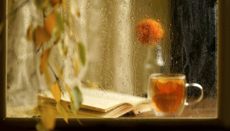 a cup of fragrant tea with an open book on a wooden windowsill against the background of an autumn rainy windowの写真素材