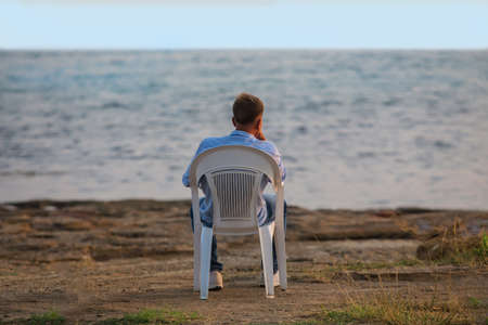 young man sits in a plastic chair by the sea in the evening at sunsetの写真素材