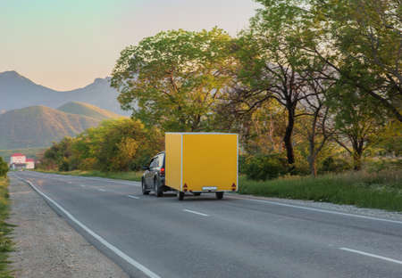 Car with Trailer moves along the road to the mountains at sunsetの写真素材