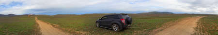 Panorama with SUV and dirt road in the field leading to the mountains
