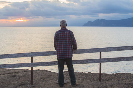 Old european man stands by the fence and looks at the sunset over the seaの写真素材