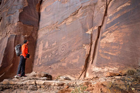 Man looking at rock art of Pueblo Indians. Owl, thunderbird, goat and a man figure petroglyphs on sandstone wall near Moab in Utah. Moab. Utah. United States.のeditorial素材
