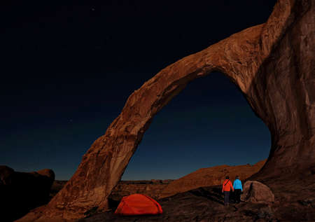 Camping tent and married couple enjoying view of natural arch and stars at night. Sandstone Corona arch in a  rock desert near Moab in Utah. The United States of America.の写真素材