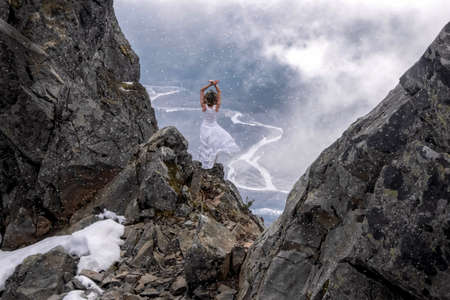 Woman in white dress standing on the edge  of a steep rock with a river view in clouds in a snow storm. First snow storm in North Cascades National Park. Concrete. Washington State. United States of America.の写真素材