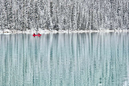 Two people canoeing in red boat on calm blue lake surrounded by winter forest covered with snow.  Beautiful reflections of winter forest and canoe in Lake Louise. Canadian Rockies. Banff National Park. Alberta. Canada.の写真素材
