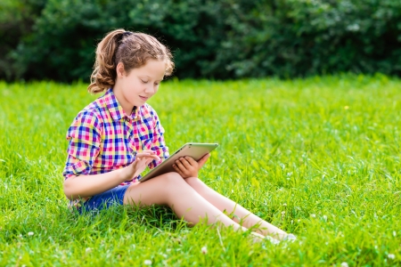 Outdoor portrait of a pretty teenager girl in casual clothes sitting on the grass with digital tablet on her knees, reading and surfingの写真素材