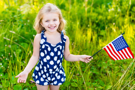 Smiling little girl with long curly blond hair holding american flag and waving it outdoor portrait on sunny day in summer park. Independence Day Flag Day conceptの写真素材