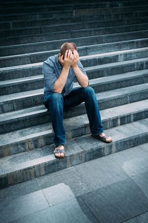 Outdoor portrait of despaired young man covering his face with hands sitting on stairs. Feelings of sadness, despair and tragedyの写真素材