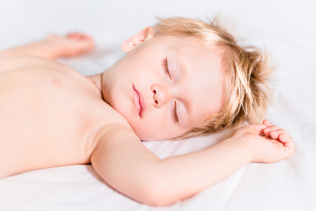 Close-up portrait of cute little kid with blond hair sleeping on white bad. Carefree childhood conceptの写真素材