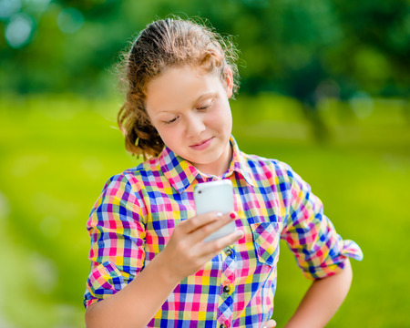 Pretty smiling teenage girl in casual clothes with smartphone in her hand, looking at screen, reading a message    の写真素材