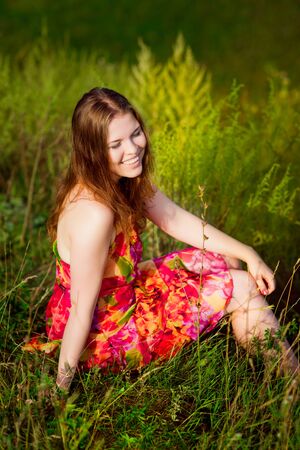 Redhaired young woman with long ginger hair and closed eyes sitting on grass and smiling on hot sunny day. Carefree freedom and leisure conceptの写真素材