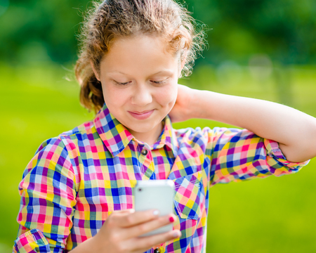 Beautiful smiling teenage girl in casual clothes with smartphone in her hand looking at screen reading a message  in sunny day in summer parkの写真素材