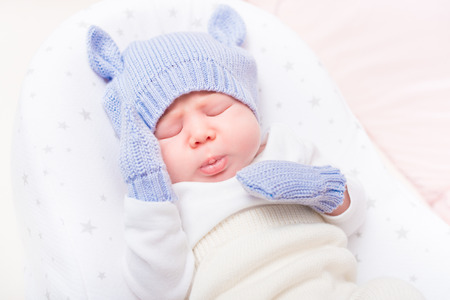 Sweet little baby wearing knitted blue hat with ears and mittens lying in beautiful cradle with closed eyes and making sad face. Security and childcare conceptの写真素材