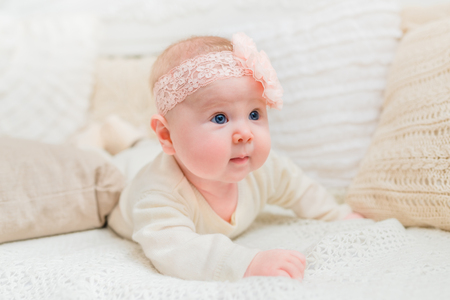 Amazed cute little baby with chubby cheeks wearing white clothes and pink band with flower lying on bed with knitted pillows. Babyhood and childhood conceptの写真素材