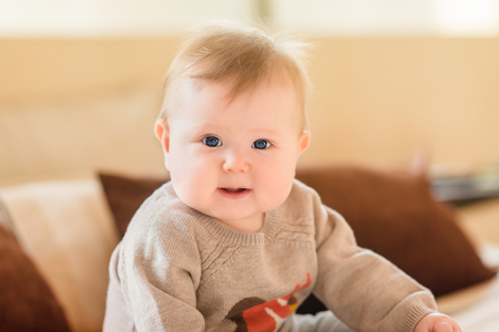 Portrait of smiling little child with blond hair and blue eyes wearing knitted sweater sitting on sofa and looking at camera. Happy childhoodの写真素材