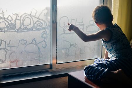 Back view of little child handrawning on fogged glass looking through the window, closeupの写真素材