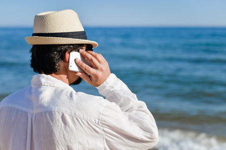 Back view closeup of person talking on cell phone walking by open water blue sky horizon, sunlight outdoors background.の写真素材