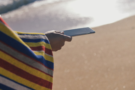 Person hand hold mobile phone with beautiful beach in the background, closeupの写真素材
