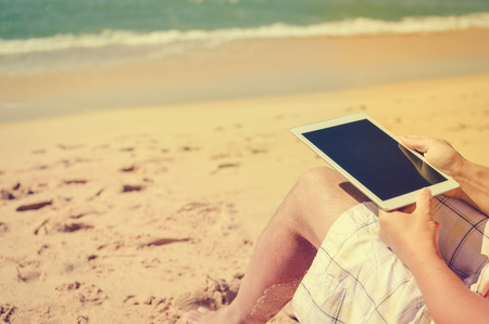 Back view of business man sitting and working with tablet computer on sunny beach outdoors...