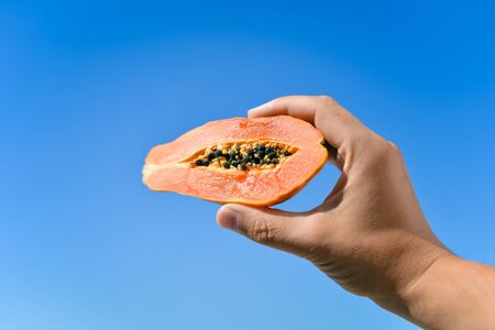 Close up on person hand holding a half papaya fruit on sunny blue sky background