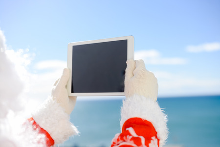 Santa Claus holding choosing receivers of gifts on tablet computer on sunny day blue sky outdoors background. Back side view closeup imageの写真素材