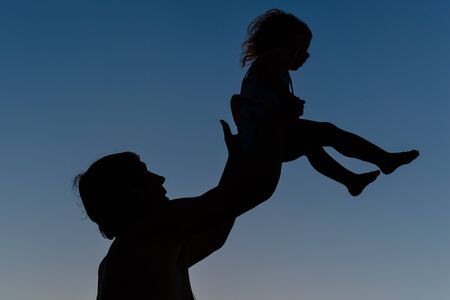 Silhouette of happy family, joyful father throwing up his child in the park on the sunset sky...