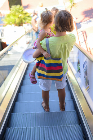 Back view of beautiful toddler girl walk with mother, escalator stairs background, closeup view. Moving staircase in shopping centre, office buildingの写真素材