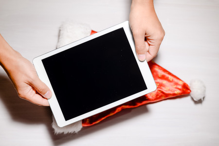 Top view of hand holding tablet computer and Santa Claus hat on light table backgroundの写真素材