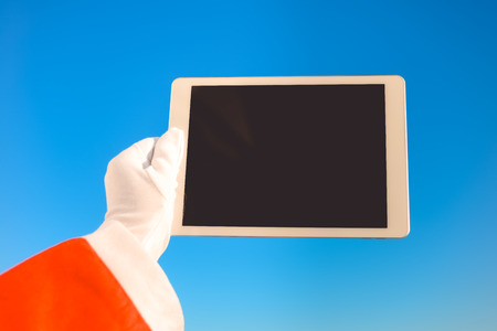 Santa Claus holding using mobile smartphone tablet pc ready for Christmas time on sunny blue sky outdoors backgroundの写真素材