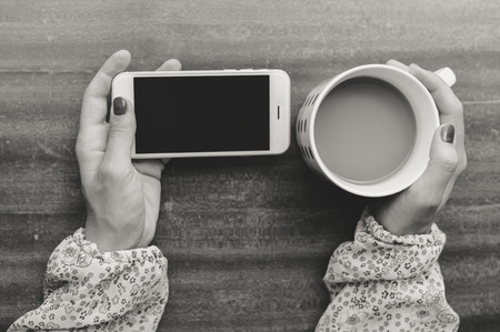 Top view on hand using smartphone and holding cup of coffee, wooden table background