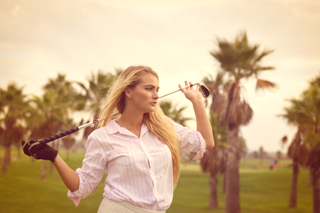 Fashion sport style beautiful woman standing holding golf club on outdoors green field backgroundの写真素材