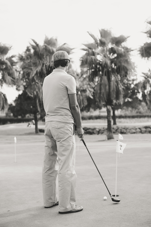 Back view of active man holding playing vintage wooden golf club on palm tree grass field outdoors...